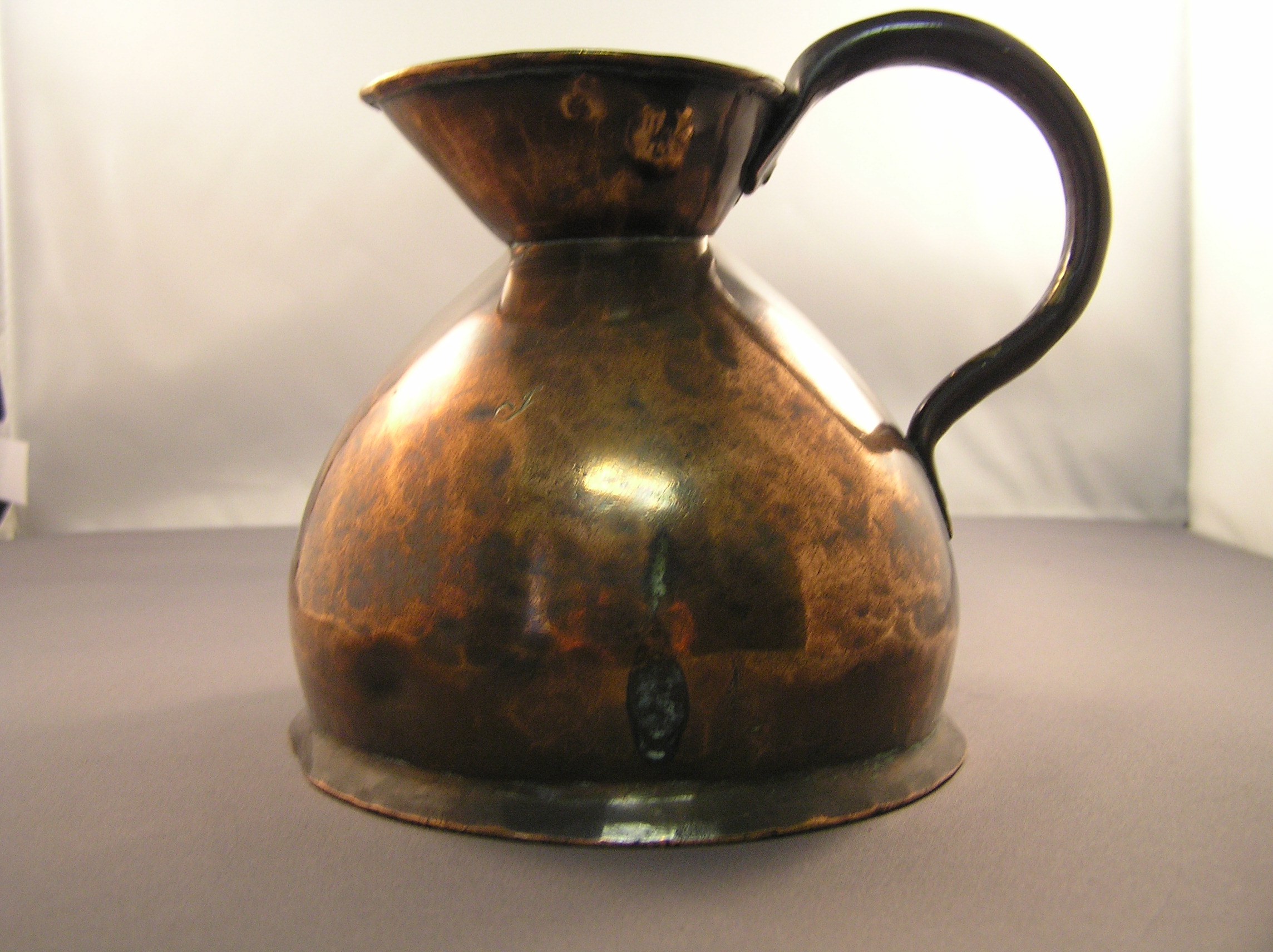 A copper measure [volume not marked] with a flat base.  The sides taper up to a wide-lipped mouth.  A rolled copper handle is riveted onto this lip and to the side of the jug. Stamped inside the rim - 3 Government inspection stamps [4 crowns above B.T]